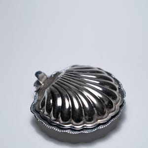 Può includere: Un piatto argentato a forma di conchiglia, con un design a coste e un bordo perlato. Il piatto ha una piccola maniglia e poggia su una superficie bianca. Questo pezzo decorativo viene probabilmente utilizzato per servire o esporre.