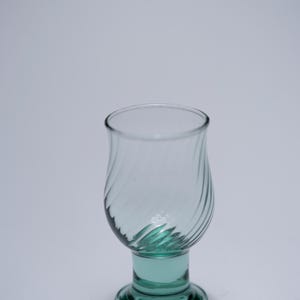 Può includere: Un bicchiere trasparente con base e stelo color verde acqua. Il bicchiere ha una forma a tulipano con un motivo a spirale sulla coppa. La base è rotonda e lo stelo è corto. Probabilmente viene utilizzato per servire bevande.
