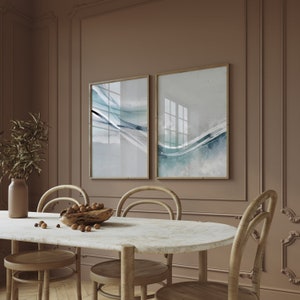 May include: Two framed abstract art prints with gold frames hang on a brown wall. The art features blue and gray watercolor-style designs. A dining table with a marble top and wooden chairs is in the foreground.