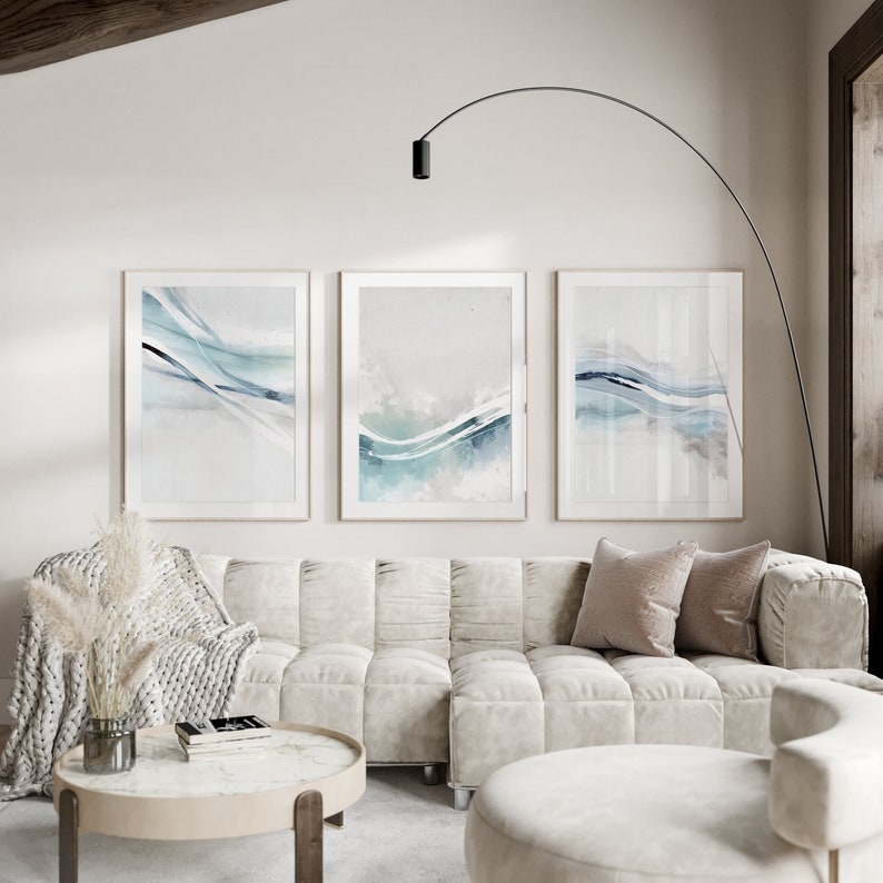 May include: Three framed abstract art prints with light blue and teal watercolor wave designs. The artwork hangs above a cream-colored tufted sofa with decorative pillows. A round coffee table and a floor lamp complete the living room setting.