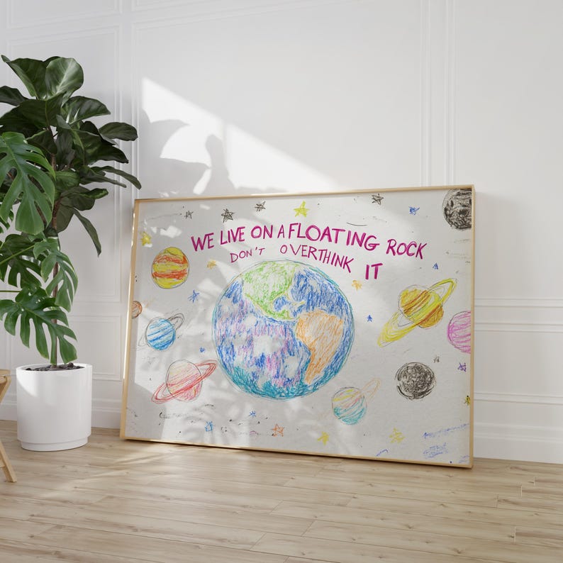 May include: A framed print with a white background and colorful crayon drawings of planets and stars. The text "WE LIVE ON A FLOATING ROCK DON'T OVERTHINK IT" is written in pink.