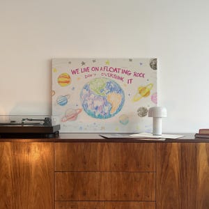May include: A white canvas with a colorful drawing of the Earth and planets with the text "WE LIVE ON A FLOATING ROCK DON'T OVERTHINK IT" in pink and white. The canvas is hanging on a white wall above a wooden cabinet with a record player and a white lamp.