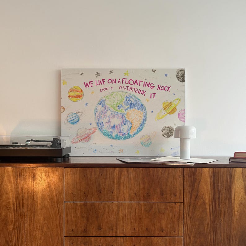 May include: A white canvas with a colorful drawing of the Earth and planets with the text "WE LIVE ON A FLOATING ROCK DON'T OVERTHINK IT" in pink and white. The canvas is hanging on a white wall above a wooden cabinet with a record player and a white lamp.