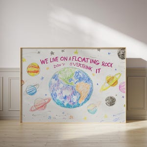 May include: A white framed print with a colorful drawing of the Earth and planets. The text reads "We live on a floating rock don't overthink it".