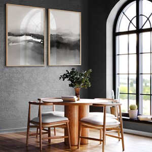 May include: A round wooden dining table with four chairs. The table has a natural wood finish and the chairs have white cushions. The table is set with a vase of greenery and a stack of books. The room has a dark grey wall and a large arched window with black trim.