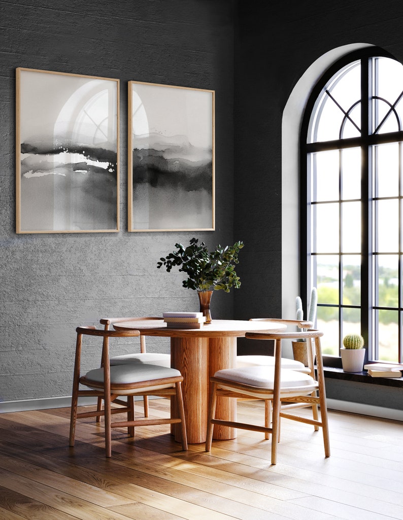 May include: A round wooden dining table with four chairs. The table has a natural wood finish and the chairs have white cushions. The table is set with a vase of greenery and a stack of books. The room has a dark grey wall and a large arched window with black trim.