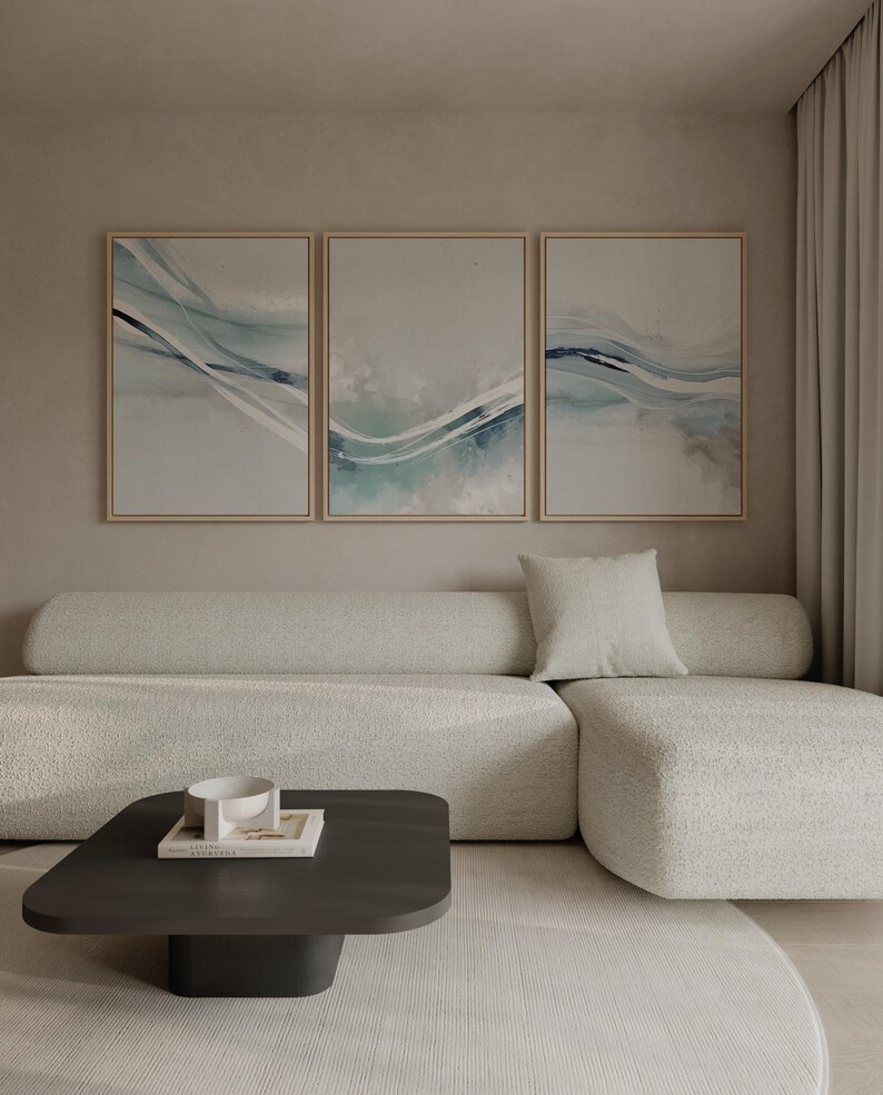 May include: Three framed abstract art pieces with flowing blue and white designs hang above a cream-colored sectional sofa. A dark brown square coffee table with a white bowl and book sits on a light-colored rug. The room has a neutral color palette.