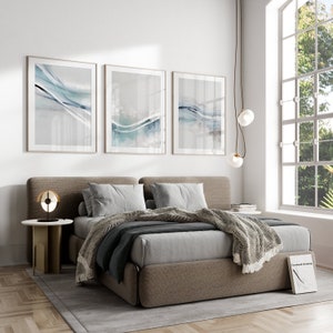 May include: A bedroom scene featuring a bed with a neutral-toned headboard, gray bedding, and a patterned throw blanket. Three framed abstract art pieces with blue and gray watercolor designs hang above the bed. A modern lamp and side table are on the left.