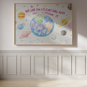 May include: A white framed poster with a colorful drawing of the Earth and planets. The text on the poster reads "We live on a floating rock. Don't overthink it."