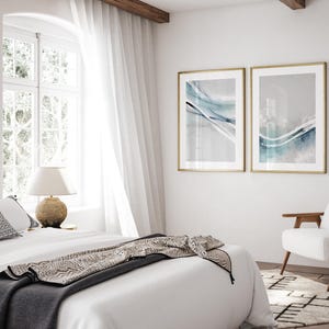 May include: Two framed abstract art prints with gold frames hang on a white wall. The prints feature flowing blue and white watercolor designs. A bed with white linens and a patterned throw is in the foreground, with a white chair and rug visible.