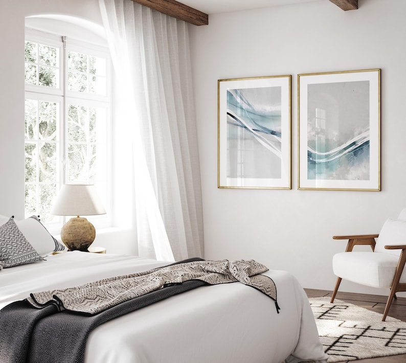 May include: Two framed abstract art prints with gold frames hang on a white wall. The prints feature flowing blue and white watercolor designs. A bed with white linens and a patterned throw is in the foreground, with a white chair and rug visible.