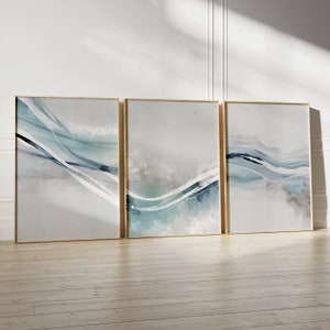 May include: Three framed abstract art prints. Each features a gold frame and a watercolor design with flowing lines in shades of blue, teal, and gray. The artwork is displayed against a white wall and wooden floor.