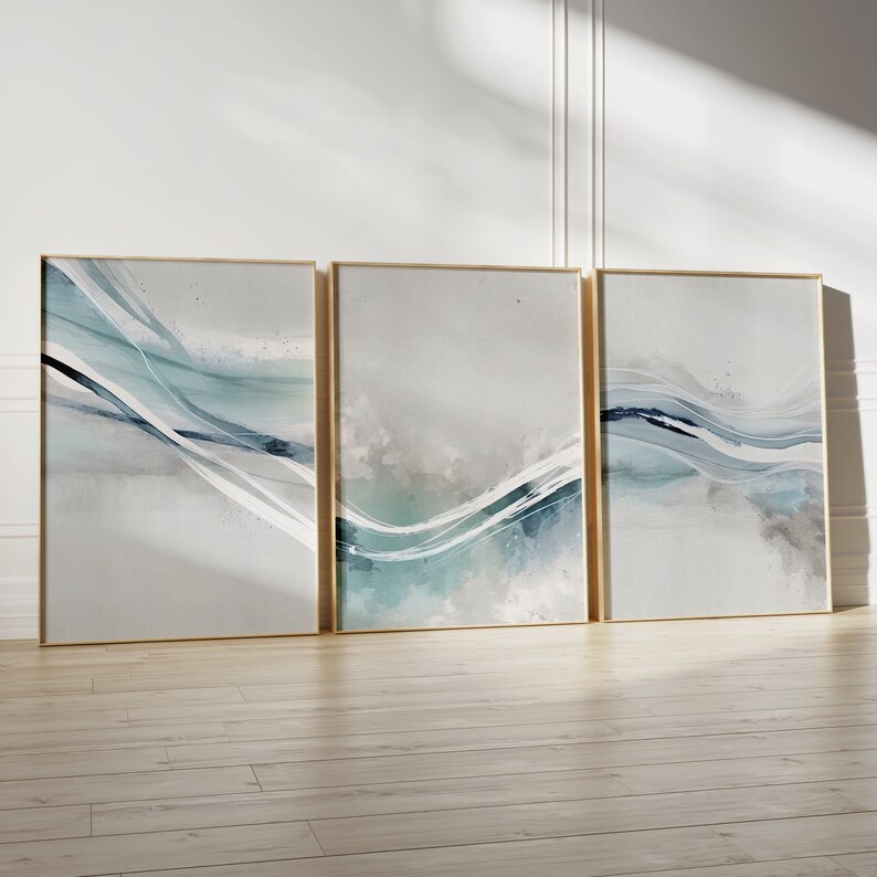 May include: Three framed abstract art prints. Each features a gold frame and a watercolor design with flowing lines in shades of blue, teal, and gray. The artwork is displayed against a white wall and wooden floor.