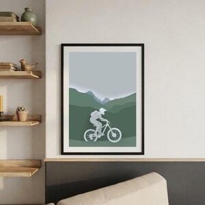 Può includere: Una stampa artistica da parete incorniciata che mostra la sagoma grigio chiaro di una persona in sella a una mountain bike. Lo sfondo presenta colline e montagne verdi stratificate sotto un cielo grigio pallido, creando profondità.