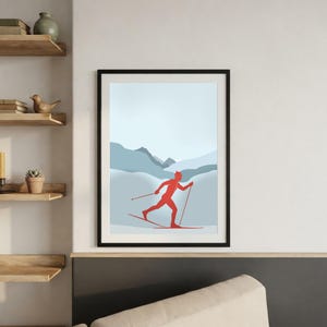 Può includere: Una stampa artistica incorniciata che raffigura la sagoma rossa di uno sciatore di fondo in movimento su uno sfondo di montagne stratificate blu chiaro e grigie sotto un cielo pallido. L'opera è racchiusa in una semplice cornice nera.