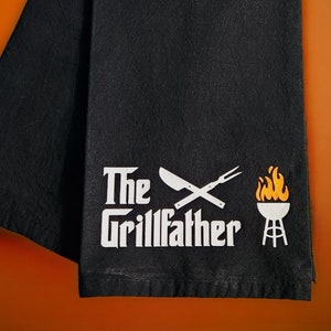May include: Black kitchen towel with the text "The Grillfather" in white, along with a knife, fork, grill, and flames graphic. The towel is folded and displayed against an orange background.