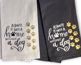 May include: Two folded kitchen towels, one cream and one dark gray. The cream towel has gold paw prints and the text "A HOUSE IS NOT A HOME WITHOUT A DOG" in black. The dark gray towel has gold paw prints and white text.