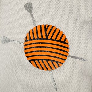May include: An orange and black yarn ball with silver knitting needles on a light gray background. The yarn ball has horizontal and diagonal black stripes. The knitting needles are silver and positioned around the yarn ball.