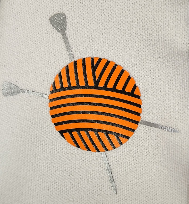 May include: An orange and black yarn ball with silver knitting needles on a light gray background. The yarn ball has horizontal and diagonal black stripes. The knitting needles are silver and positioned around the yarn ball.
