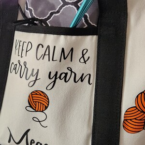 May include: Cream-colored tote bag with black trim and handles, featuring the text "Keep Calm & Carry Yarn." The bag has a front pocket and is decorated with orange yarn ball graphics. Knitting needles are visible in the pocket.