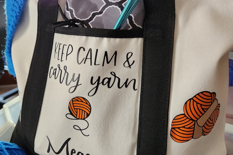 May include: Cream-colored tote bag with black trim and handles, featuring the text "Keep Calm & Carry Yarn." The bag has a front pocket and is decorated with orange yarn ball graphics. Knitting needles are visible in the pocket.