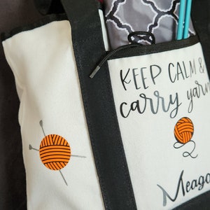 May include: A white and black tote bag with the text "KEEP CALM & carry yarn" and a yarn ball graphic. The bag has a black handle and trim, and contains knitting needles and a patterned pouch.