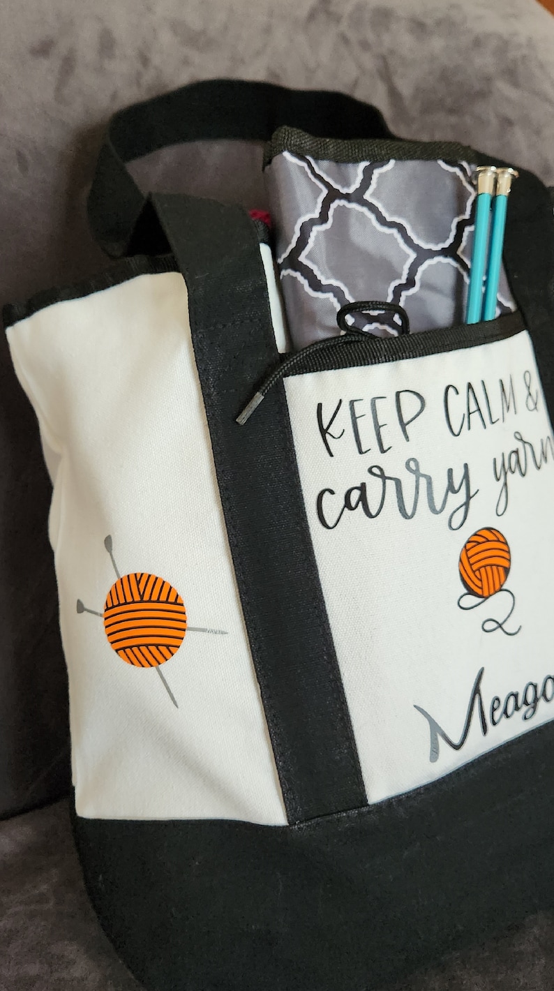 May include: A white and black tote bag with the text "KEEP CALM & carry yarn" and a yarn ball graphic. The bag has a black handle and trim, and contains knitting needles and a patterned pouch.