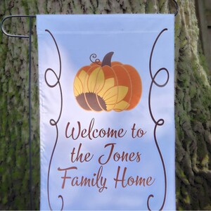 May include: A white rectangular garden flag featuring a central graphic of an orange pumpkin with yellow sunflower petals. The text reads Welcome to the Jones Family Home in brown script. The design is framed by two brown decorative scroll lines.