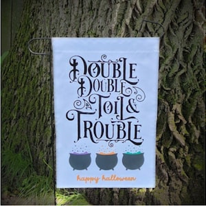 以下が含まれることがあります： Double Double Toil & Troubleと書かれた黒い装飾文字が特徴の白い長方形のガーデンフラッグ。テキストの下には、紫、オレンジ、緑の泡立つ液体が入った3つのダークグレーの釜が描かれています。happy halloweenという言葉はオレンジ色で書かれています。