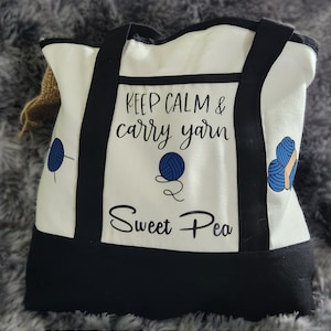 May include: A white tote bag with black accents and handles. The bag features the text "KEEP CALM & carry yarn Sweet Pea" with blue yarn ball graphics. The bag is designed for carrying yarn and craft supplies.
