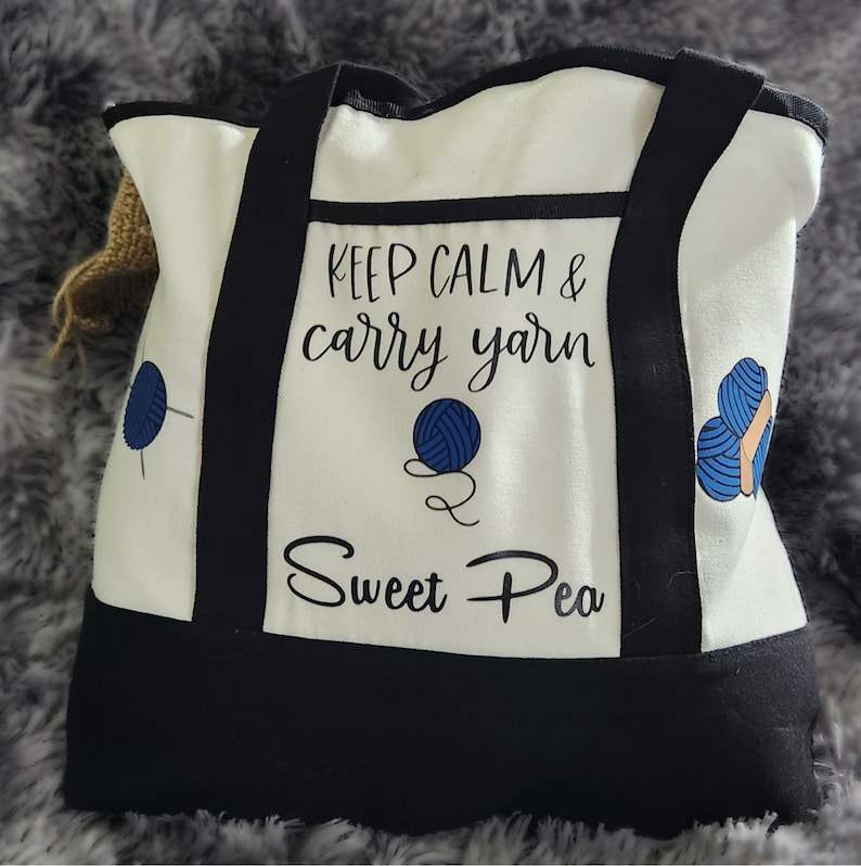 May include: A white tote bag with black accents and handles. The bag features the text "KEEP CALM & carry yarn Sweet Pea" with blue yarn ball graphics. The bag is designed for carrying yarn and craft supplies.