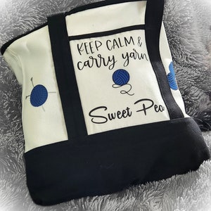 May include: Cream-colored tote bag with black trim and handles. The bag features the text "KEEP CALM & carry yarn Sweet Pea" along with blue yarn ball graphics. The bag is designed for carrying yarn and knitting supplies.