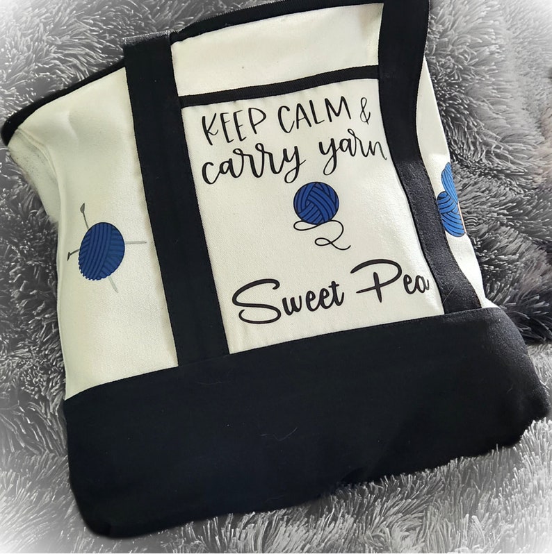 May include: Cream-colored tote bag with black trim and handles. The bag features the text "KEEP CALM & carry yarn Sweet Pea" along with blue yarn ball graphics. The bag is designed for carrying yarn and knitting supplies.