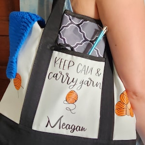 May include: A black and white tote bag with the text "KEEP CALM & carry yarn" and the name "Meagan". The bag has a pocket with a gray and white patterned case and a blue knitting needle. A blue knitted item is in the bag.