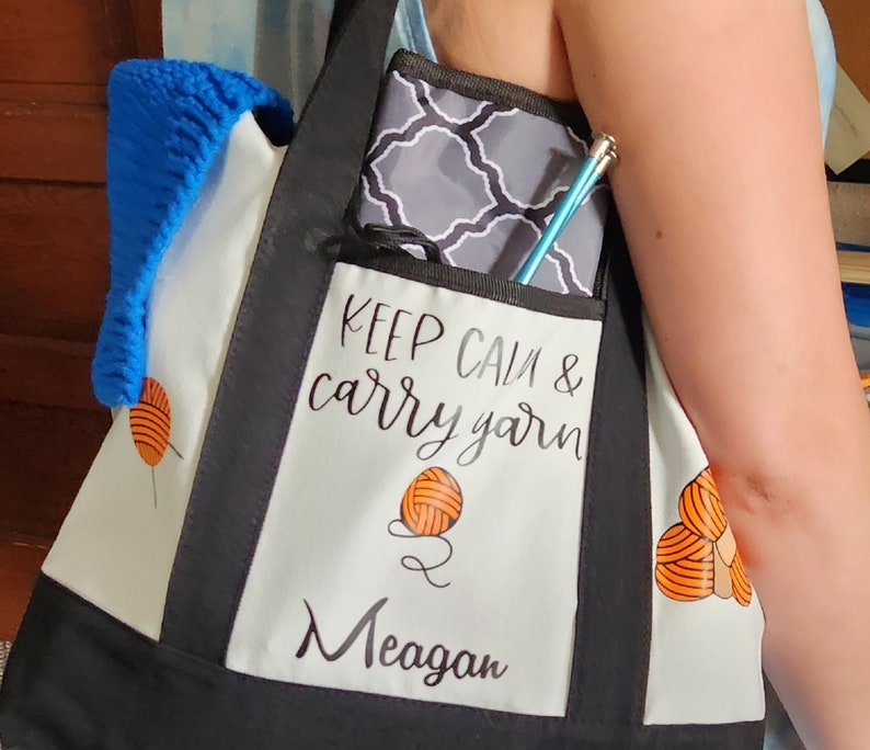 May include: A black and white tote bag with the text "KEEP CALM & carry yarn" and the name "Meagan". The bag has a pocket with a gray and white patterned case and a blue knitting needle. A blue knitted item is in the bag.