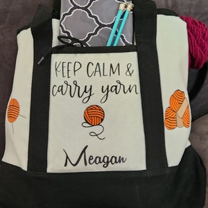May include: A black and white tote bag with the text "KEEP CALM & carry yarn" and the name "Meagan". The bag has a front pocket with a gray patterned pouch and knitting needles. A ball of pink yarn is visible.