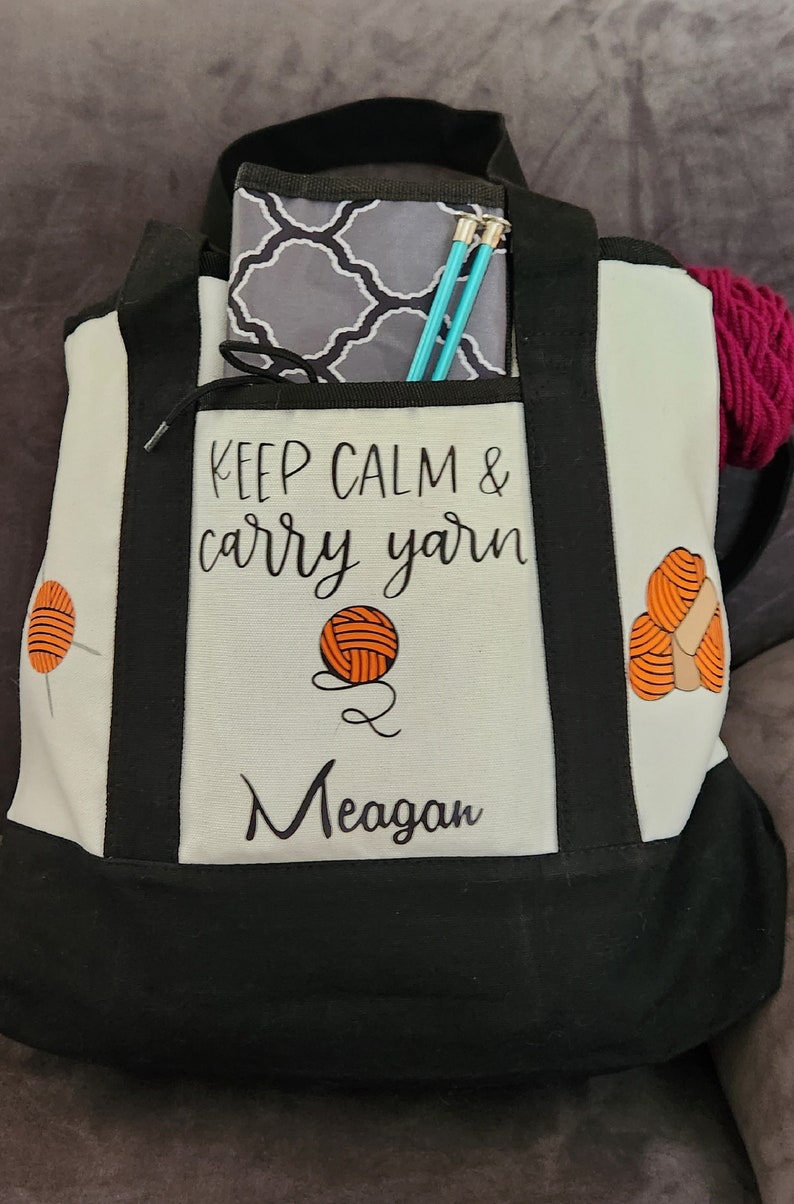 May include: A black and white tote bag with the text "KEEP CALM & carry yarn" and the name "Meagan". The bag has a front pocket with a gray patterned pouch and knitting needles. A ball of pink yarn is visible.