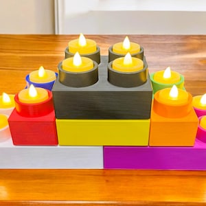 Op de afbeelding: Een kleurrijk kaarsenhouderarrangement. De opstelling bestaat uit vierkante en rechthoekige blokken in verschillende kleuren, waaronder wit, rood, geel, groen, oranje, blauw en paars. Elk blok bevat een kleine, ronde kaars met een flikkerende gele vlam.