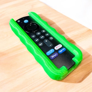 May include: A black remote control with a bright green protective cover. The remote features buttons for power, navigation, and streaming services. The green cover has a wavy design. The remote is designed for streaming services.