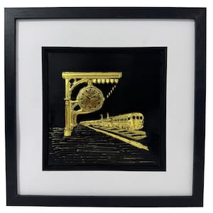 May include: Framed artwork featuring a gold-colored train station scene on a black background. The artwork includes a clock, train, and tracks, all in a metallic gold hue. The piece is encased in a black frame with a white mat.