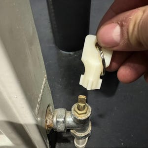 Puede incluir: Una llave de radiador de plástico blanco sostenida cerca de una válvula de latón. La llave está unida a un anillo plateado. La válvula es parte de una tubería de metal conectada a un radiador blanco.
