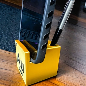 May include: A bright yellow desk organizer with the periodic table symbol "Au" for gold. A black smartphone and a silver pen are placed in the organizer. The organizer is made of a solid material and sits on a wooden surface.