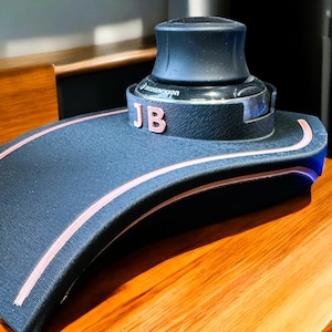 May include: A black and pink device with the initials "JB" on the side. The device has a curved, textured surface with pink trim and a cylindrical top. The device is sitting on a wooden surface.