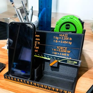 Puede incluir: Un organizador de escritorio negro con un soporte para teléfono incorporado, una regla y una cinta métrica. El organizador tiene una regla impresa a lo largo del borde frontal e incluye texto con medidas en pulgadas y libras.