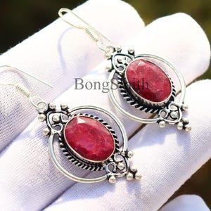 Może przedstawiać: Para srebrnych kolczyków z owalnymi, głęboko czerwonymi kamieniami. Kolczyki mają ozdobny, ażurowy wzór z dekoracyjnymi detalami wokół kamieni i prostym zapięciem na haczyk.