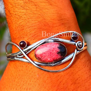 May include: A silver bracelet featuring a central oval pink and black gemstone, with two smaller red stones. The bracelet has a flowing, organic design. The text "BongSmith" is visible on the bracelet.
