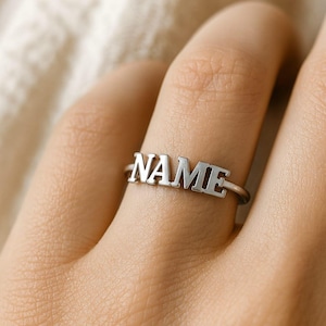 May include: A silver ring with the word "NAME" spelled out in capital letters. The ring is worn on a finger, and the letters are raised and in a sans-serif font. The ring is a simple, elegant piece of jewelry.