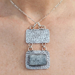 May include: A silver necklace featuring a pendant with two rectangular plates connected by jump rings. The top plate has a textured surface, while the bottom plate holds a rectangular, speckled grey and white stone in a silver bezel setting.