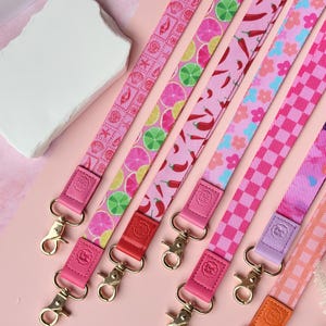 May include: A collection of colorful lanyards with various patterns, including citrus fruits, chili peppers, and floral designs. Each lanyard features a different color scheme, with pink as the primary color. The lanyards have gold-colored clasps and leather accents.
