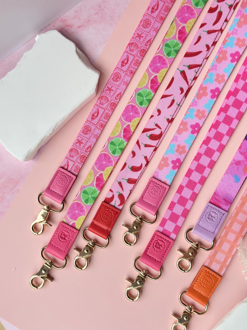 May include: A collection of colorful lanyards with various patterns, including citrus fruits, chili peppers, and floral designs. Each lanyard features a different color scheme, with pink as the primary color. The lanyards have gold-colored clasps and leather accents.